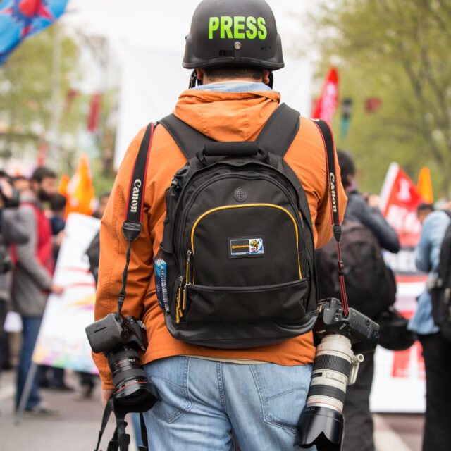 Photographer with press badge and camera gear at a crowded event.